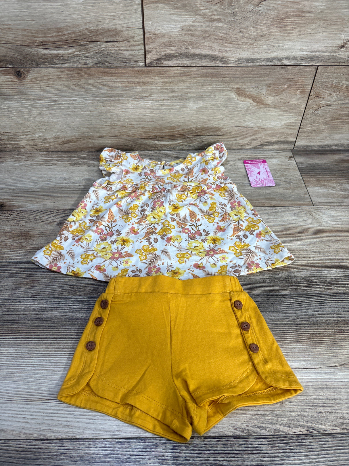 Flat lay of 2-piece baby floral flutter-sleeve top and mustard shorts, size 12 months, side button detail on wood background