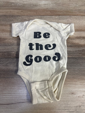 Cream baby onesie with black text 'Be the Good' on wooden surface