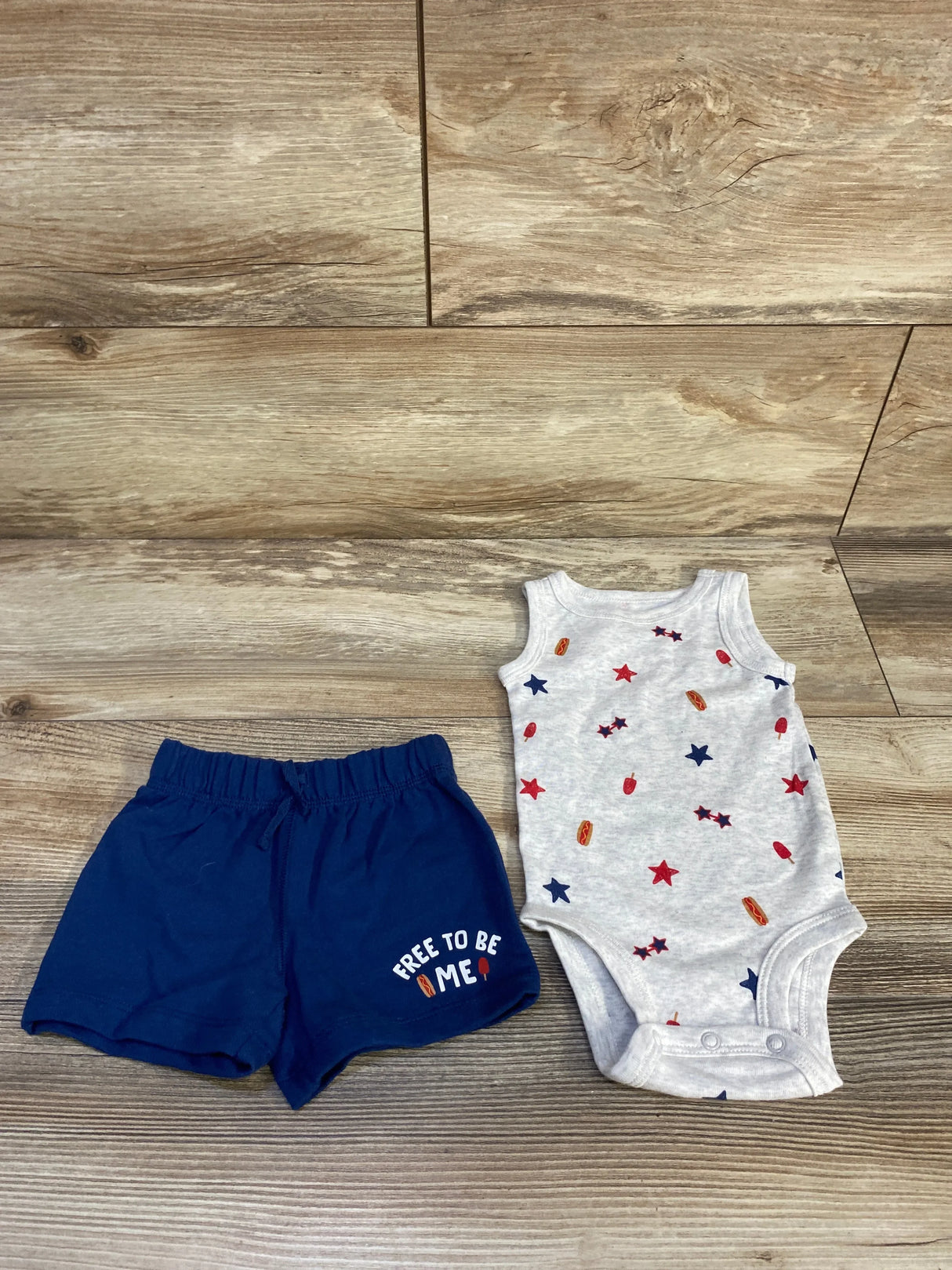 Top-down view of 2-piece baby set: grey bodysuit with star and popsicle print, navy shorts reading 'Free to be me' on wood