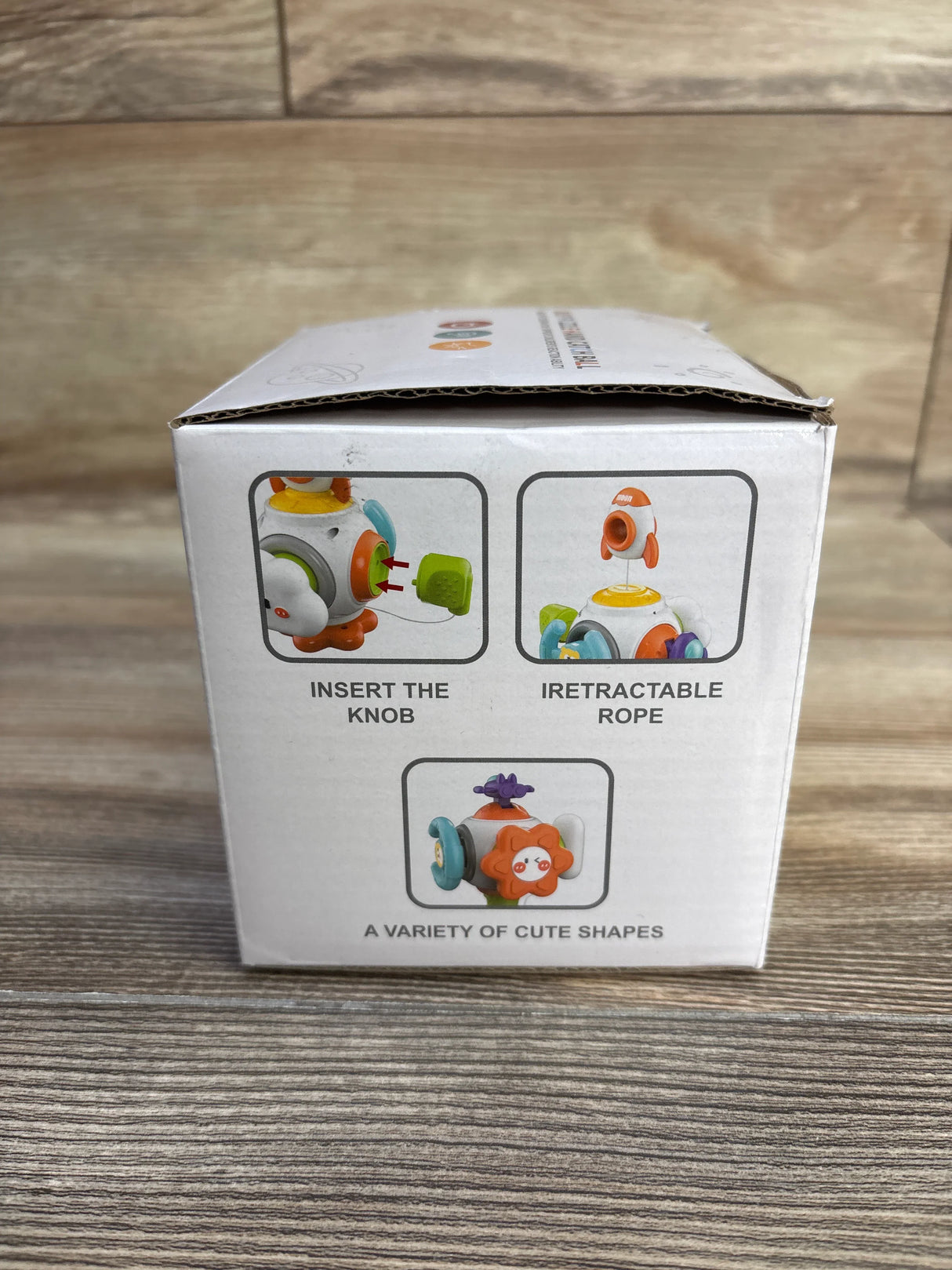 Front view of Happy Play Puzzle Busy Ball box showing toy images and the captions 'Insert the knob', 'Retractable rope', and 'A variety of cute shapes' on a wooden surface
