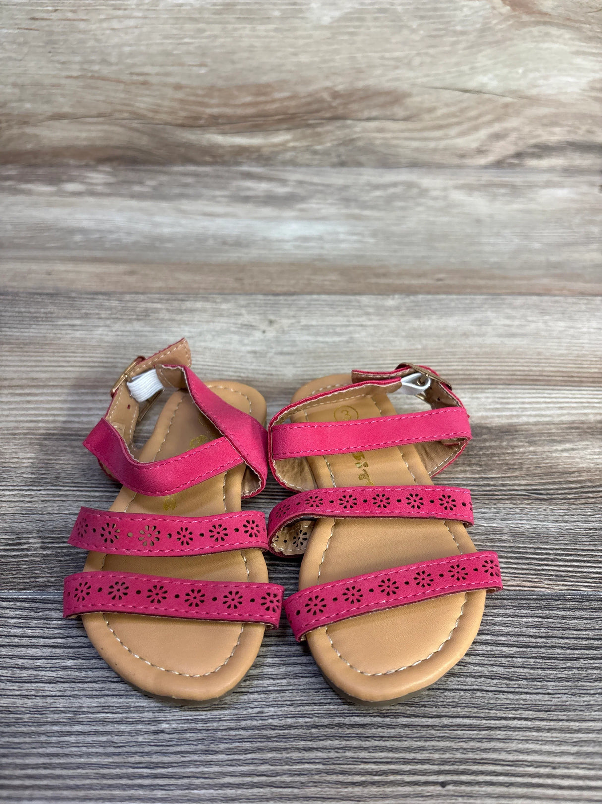 Top-down view of Ositos slingback toddler sandals showing three floral-cut pink straps and tan stitched footbed on wood background, size 5C