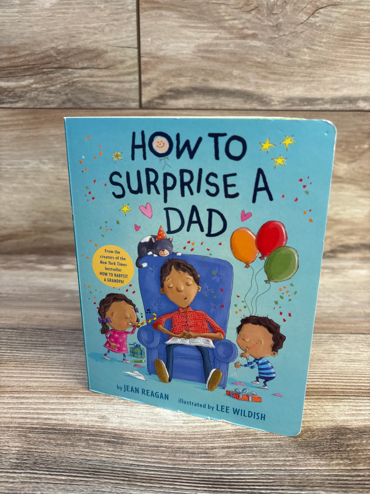 Front cover of How to Surprise a Dad board book showing dad in armchair with two children and balloons on wood background