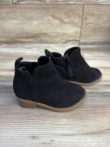 Front three-quarter view of black ankle boots showcasing side zippers and tan soles on a wood-look floor