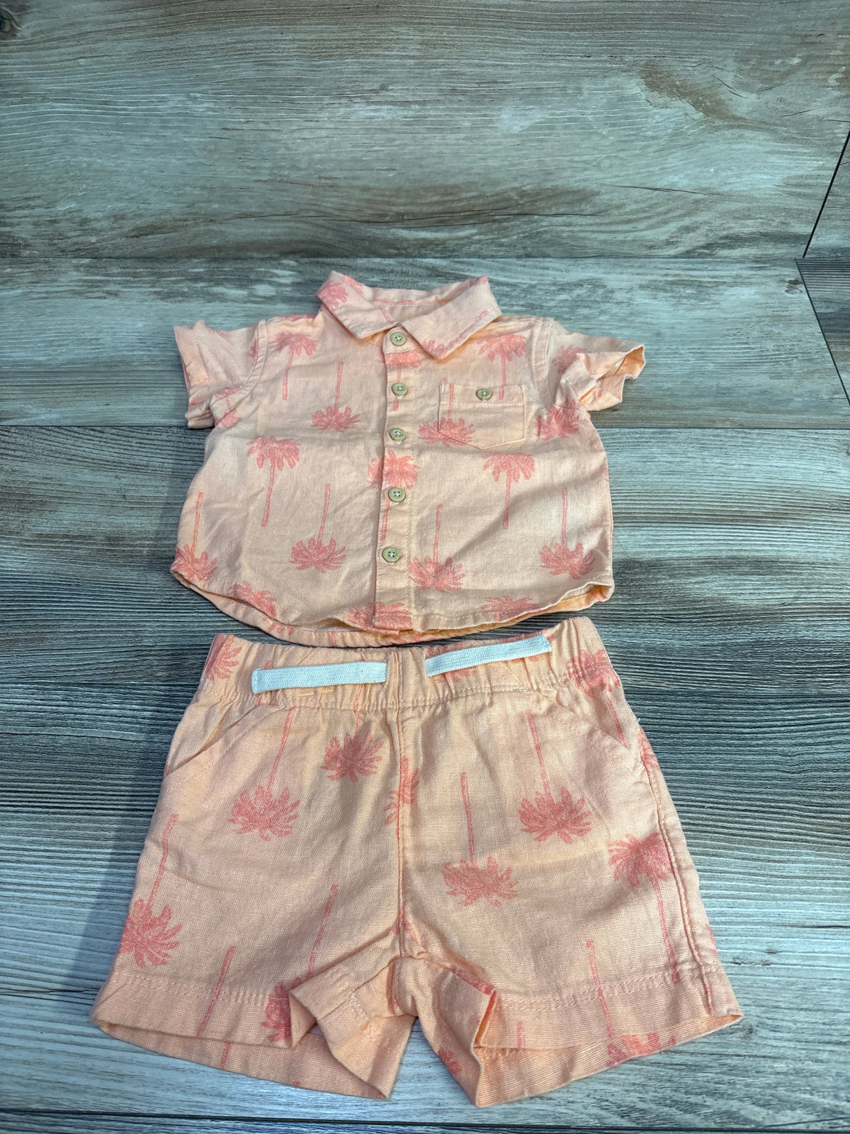 Flat lay of coral newborn two-piece button-up shirt and elastic-waist shorts with pink palm tree print on wood background