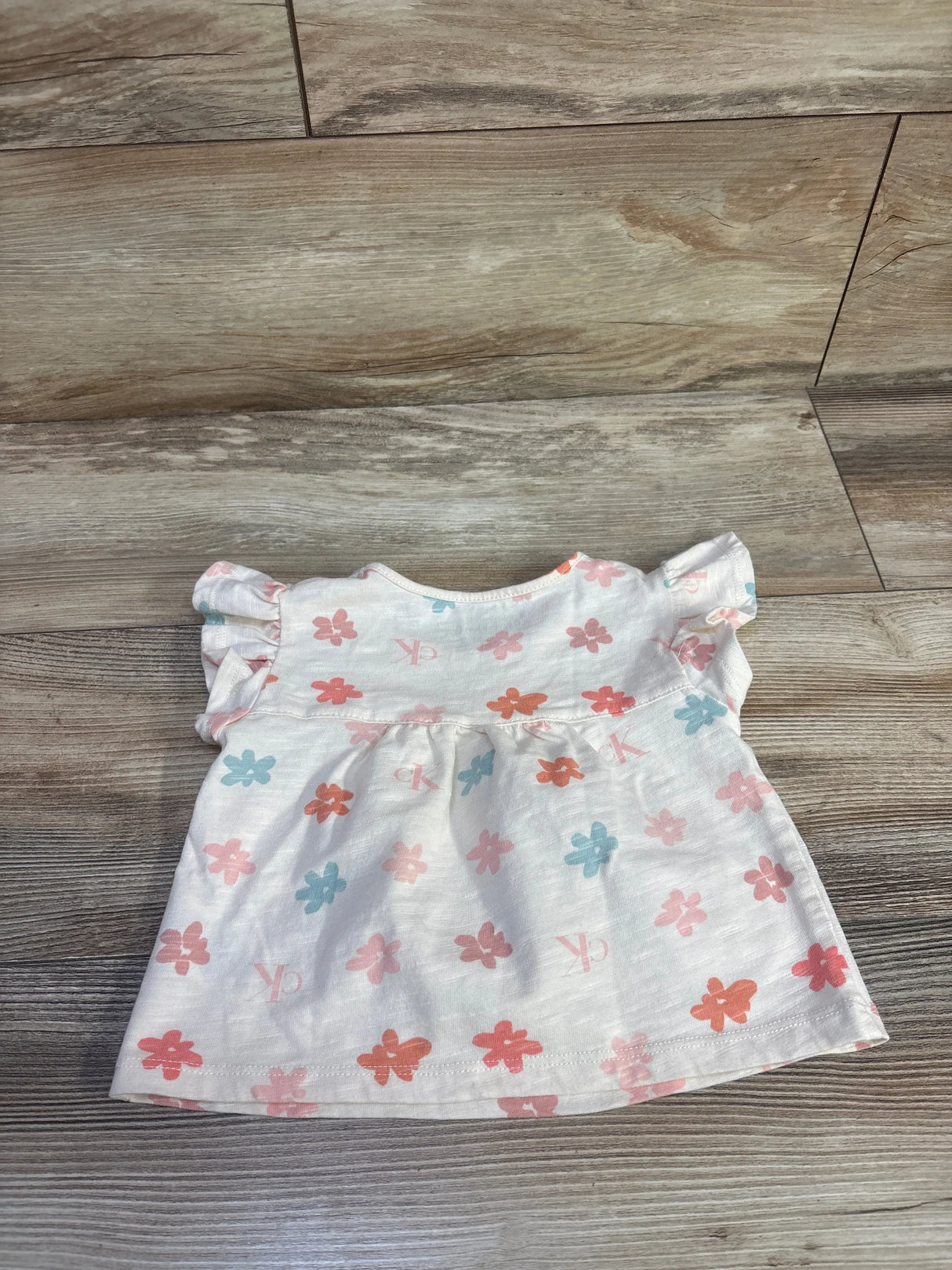 Flat lay of pink Calvin Klein toddler flutter sleeve shirt showcasing ruffled cap sleeves and pastel flower print on wood floor background