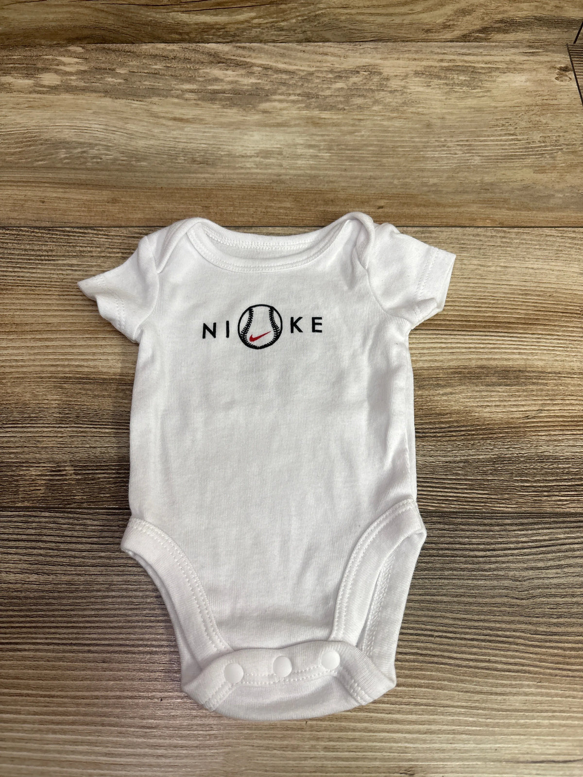 White Nike newborn bodysuit with a baseball graphic on the chest, lying on a wooden surface.