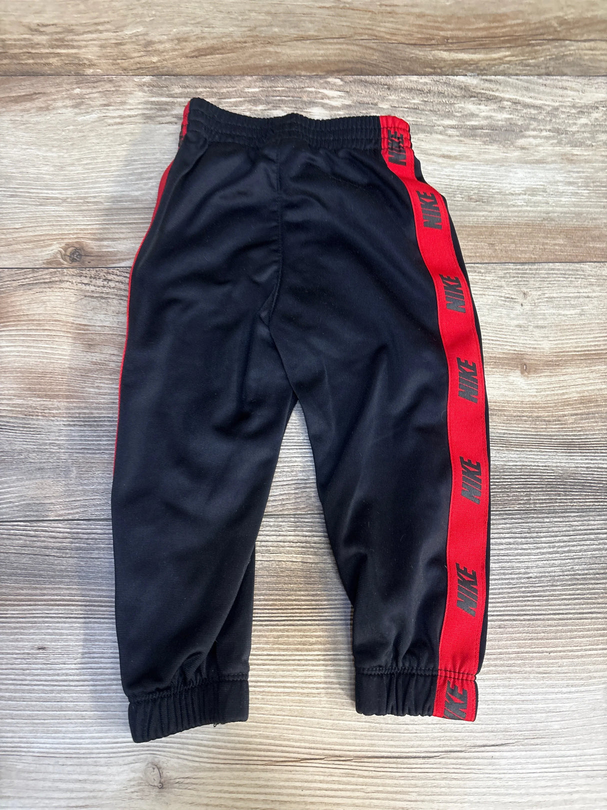 Black Nike toddler jogger pants with red side stripe and logo, laid flat on wood surface