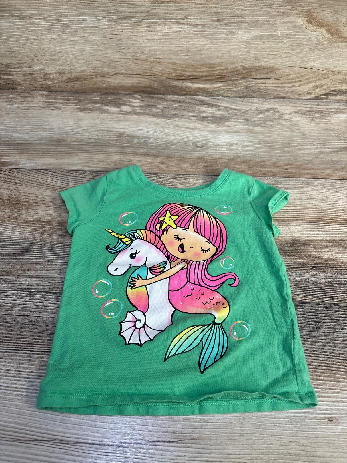Flat-lay of green Children's Place 2T t-shirt showing a pink-haired mermaid hugging a unicorn graphic, short sleeves on wood background