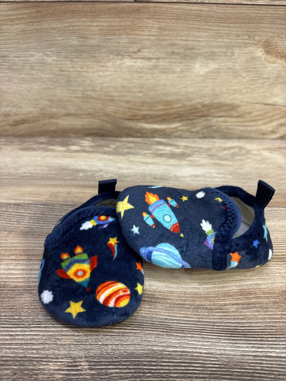 Side view of navy baby slippers size 5C showing plush fabric and colorful rocket and planet print on wood background