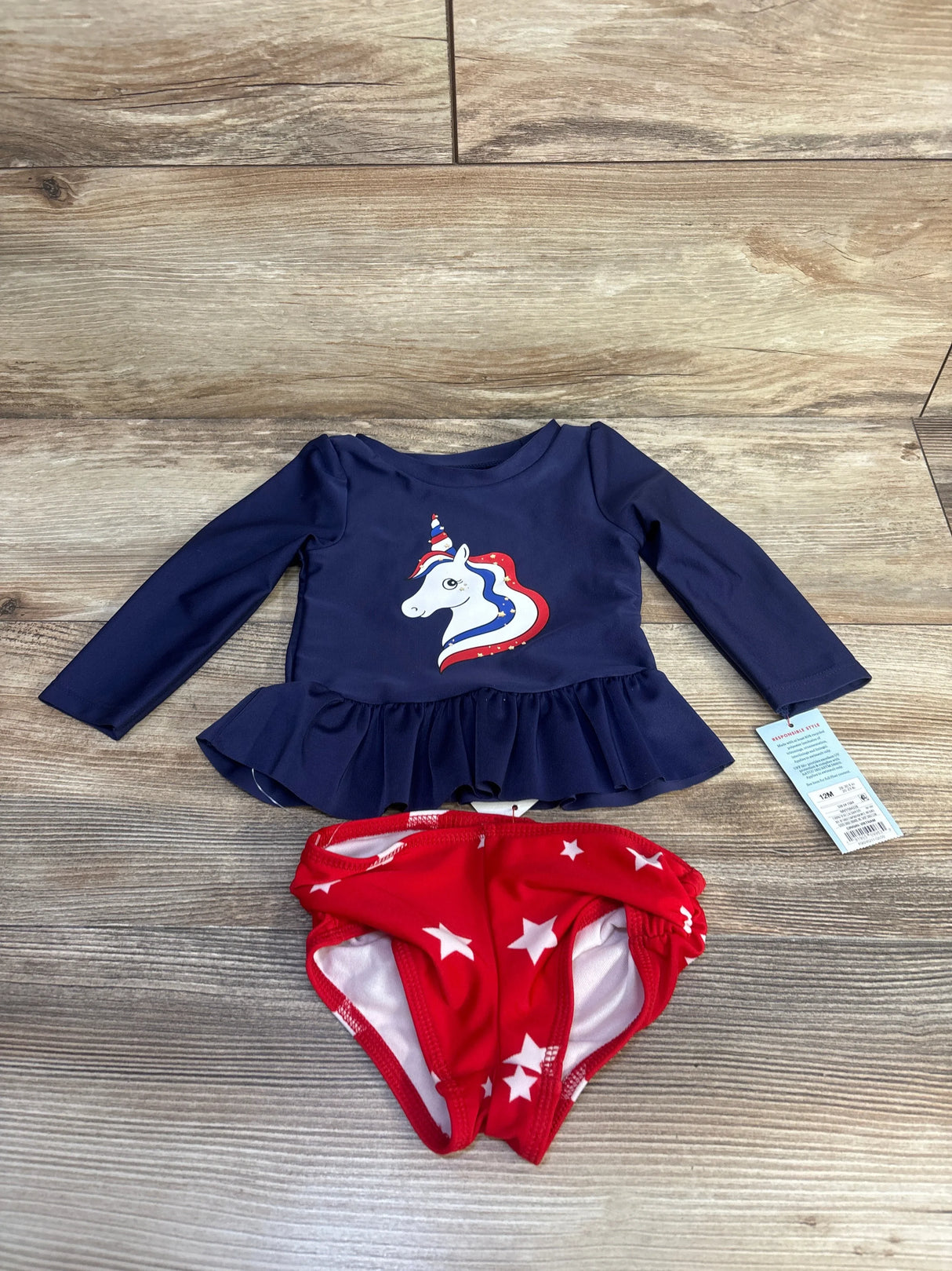 Flat lay of navy long-sleeve peplum rashguard with unicorn graphic and red star swim bottoms, size 12M on wood floor