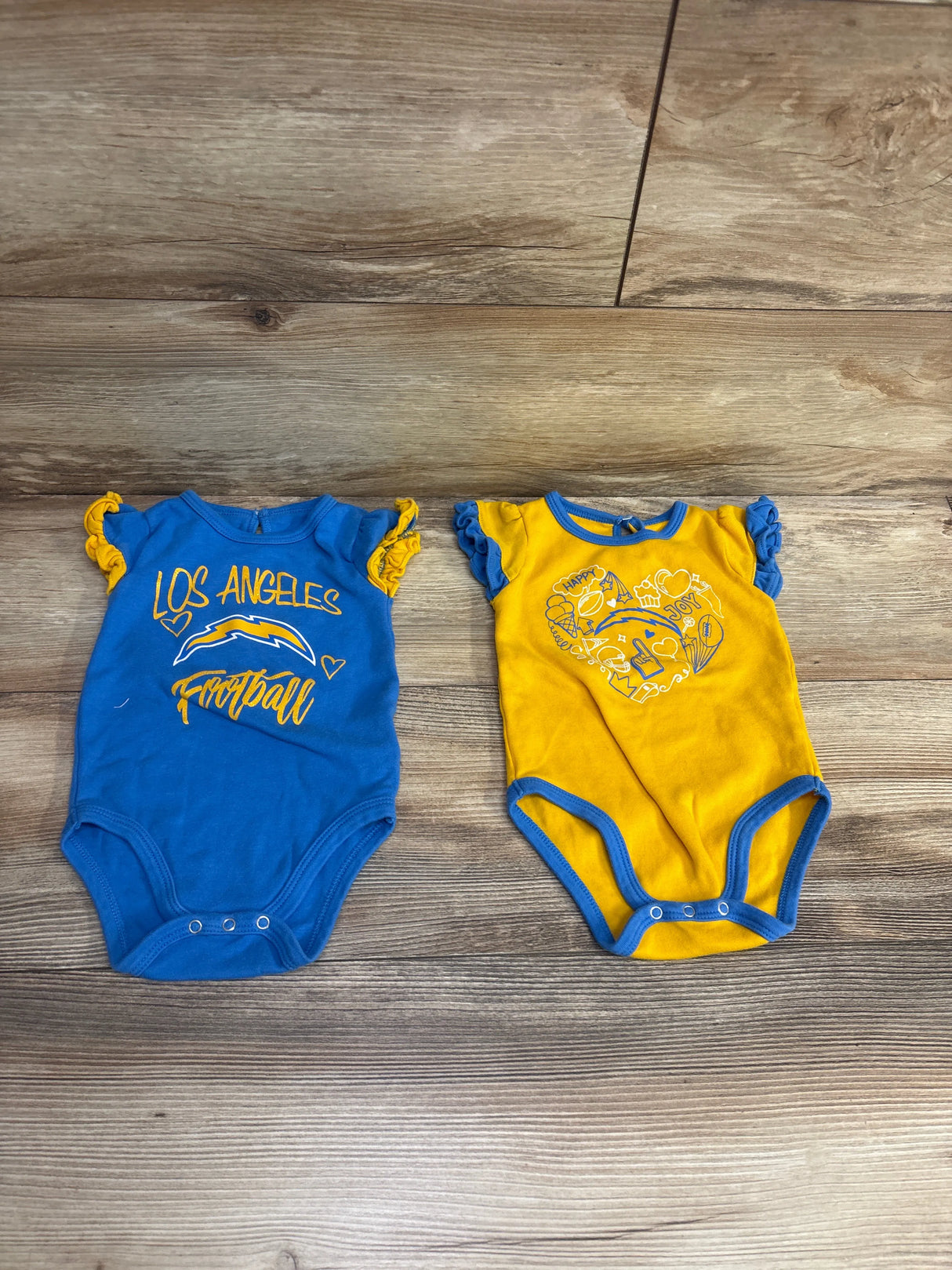 Two baby bodysuits on wooden floor; blue with Los Angeles football print, yellow with heart graphic and ruffled sleeves