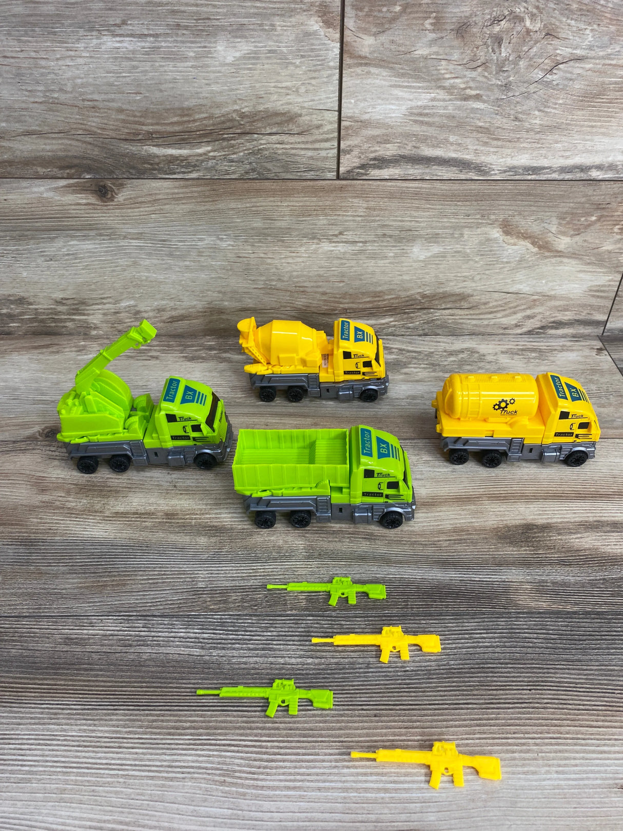Four toy construction trucks in green and yellow with six matching toy rifles on a wooden floor