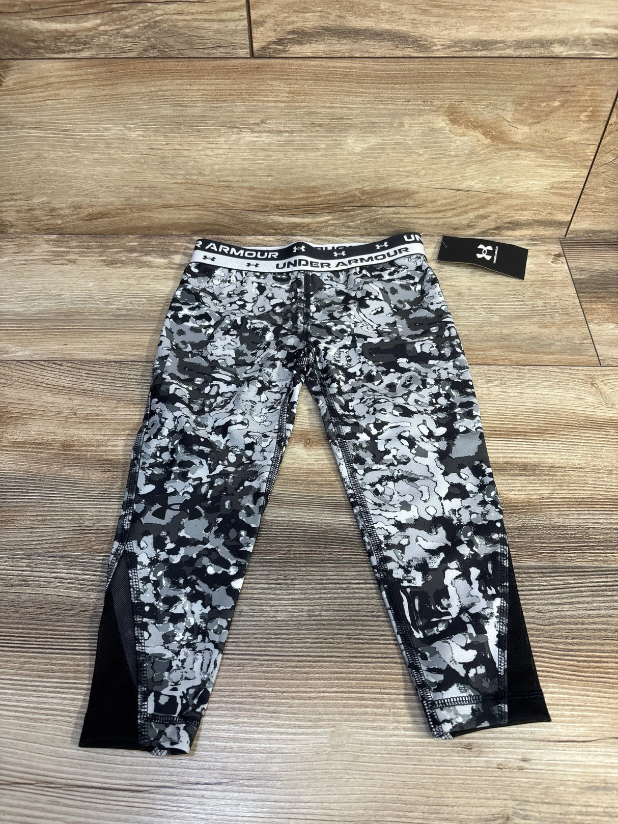 Flat-lay of Under Armour grey camo crop leggings size 5T with branded waistband and black mesh ankle panels on wood