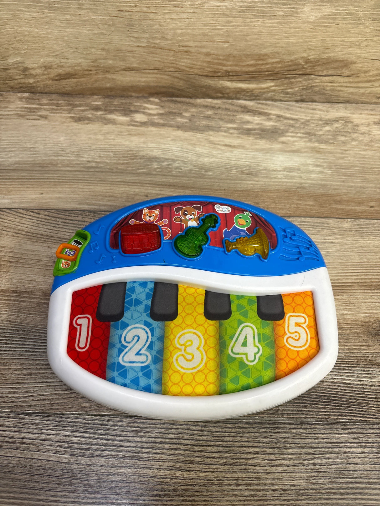 Top-down view of Baby Einstein Discover and Play Piano with five colorful numbered keys 1-5, three shaped instrument buttons on blue top, white frame on wood surface