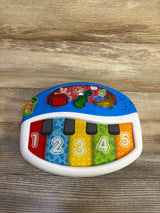 Top-down view of Baby Einstein Discover and Play Piano with five colorful numbered keys 1-5, three shaped instrument buttons on blue top, white frame on wood surface