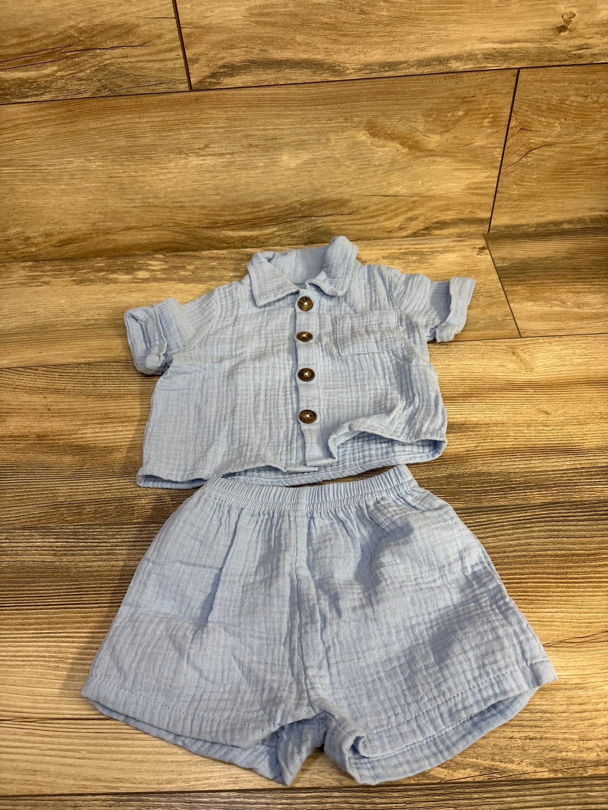 Flat lay of blue gauze baby shirt and elastic-waist shorts showing wooden buttons and crinkled texture on wood floor