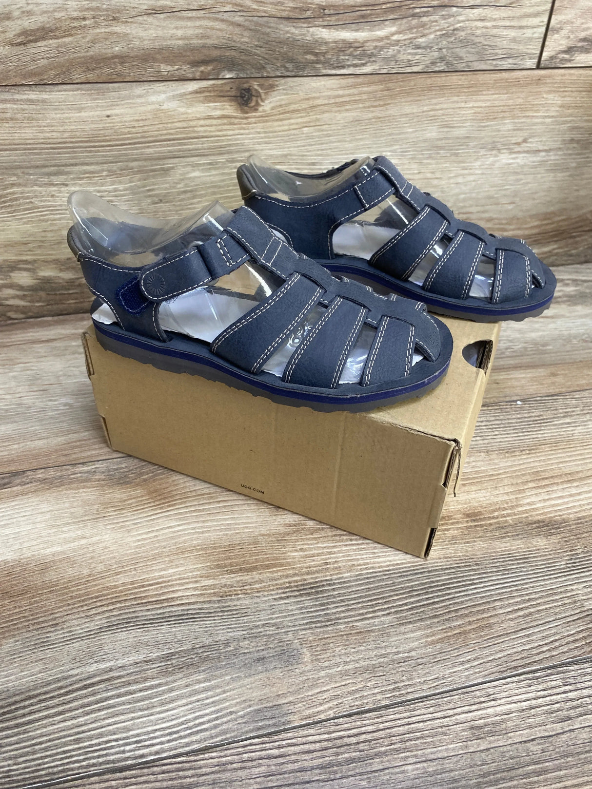 Three-quarter view of UGG Josiah kids fisherman sandal, navy, showing woven closed-toe straps and ankle Velcro, on shoebox