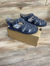 Three-quarter view of UGG Josiah kids fisherman sandal, navy, showing woven closed-toe straps and ankle Velcro, on shoebox