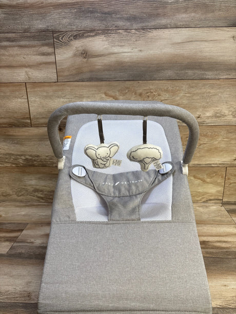 Top view of Baby Delight Highland baby bouncer in pebble grey showcasing mesh seat, adjustable safety straps, and toy bar with elephant and tree plush toys on wood floor background