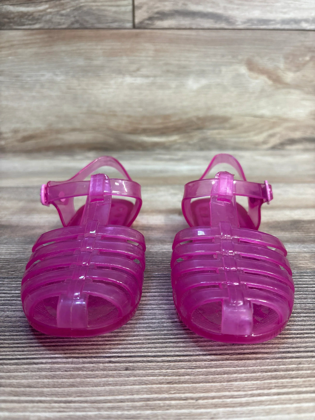 Front view of Janie & Jack Jelly Fisherman sandals showing translucent pink caged toes and ankle buckle straps on a wood background