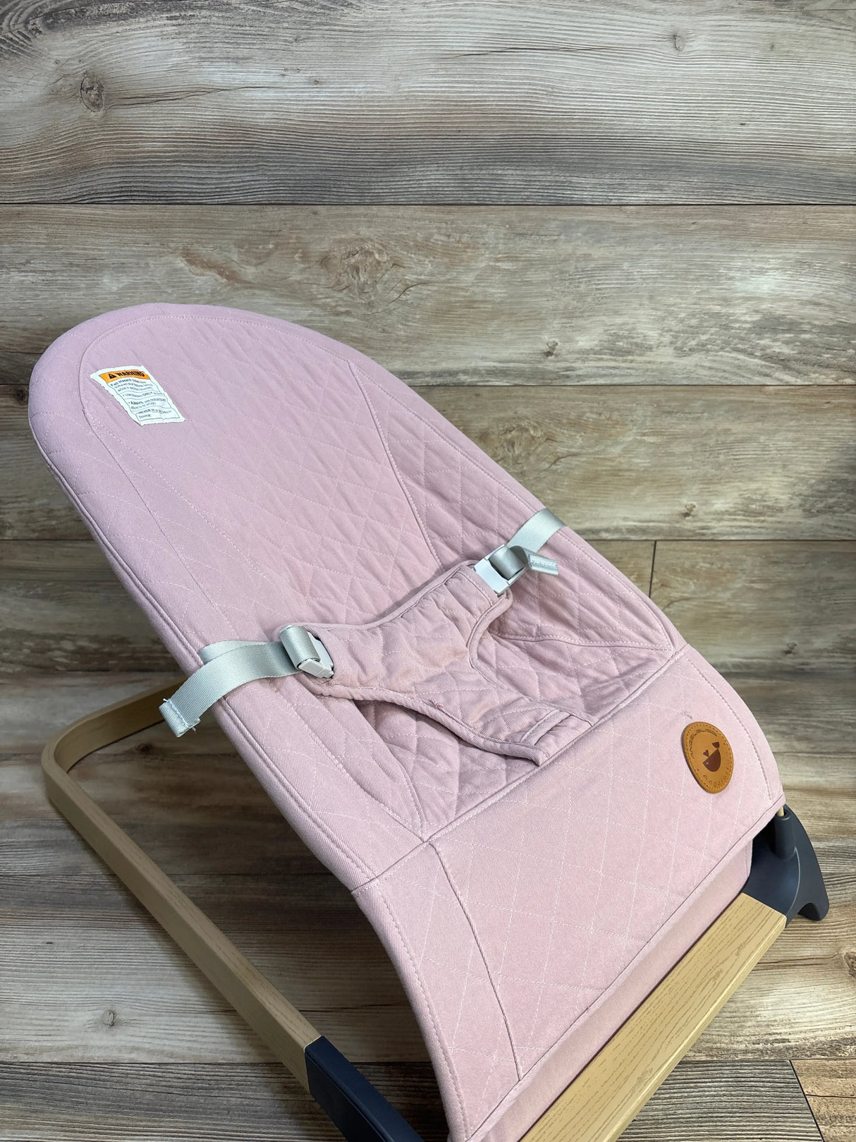 Angled view of ANGELBLISS portable baby bouncer in pink showing quilted seat, three-point harness, and wooden frame on wood background