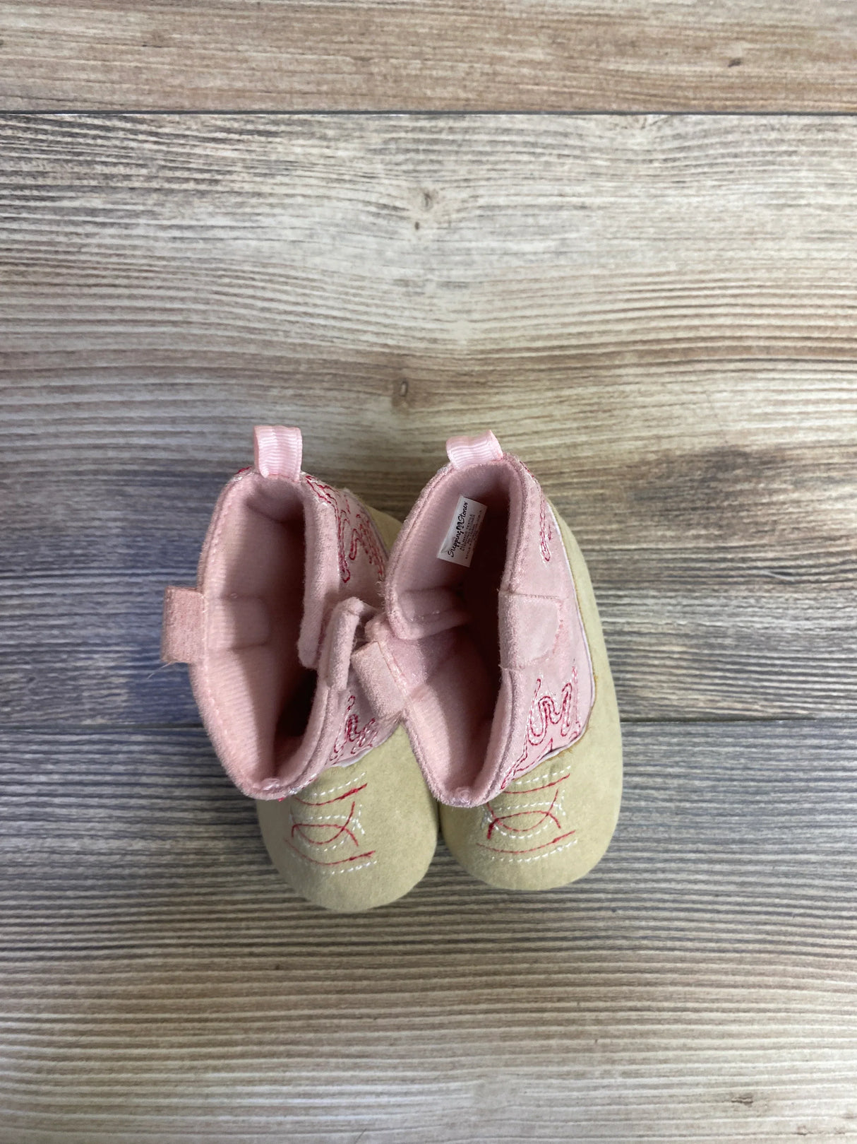 Top-down view of Stepping Stones baby cowgirl crib boots in pink showcasing embroidered cowboy stitching, pull tabs, and soft soles on wood-plank background