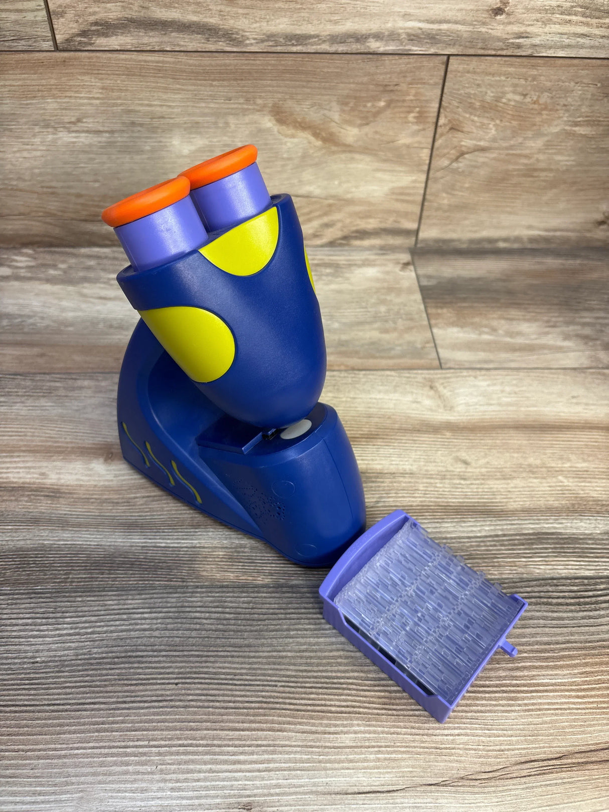Three-quarter view of GeoSafari microscope showing dual eyepieces, yellow accents and purple slide tray on wood surface