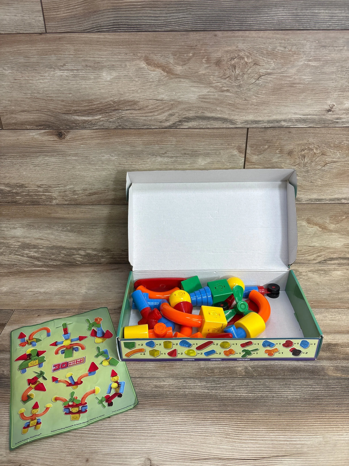 Top view of Popular Playthings Mag Builders 30pc Set showcasing colorful magnetic building pieces in original box with instruction sheet on wood plank background