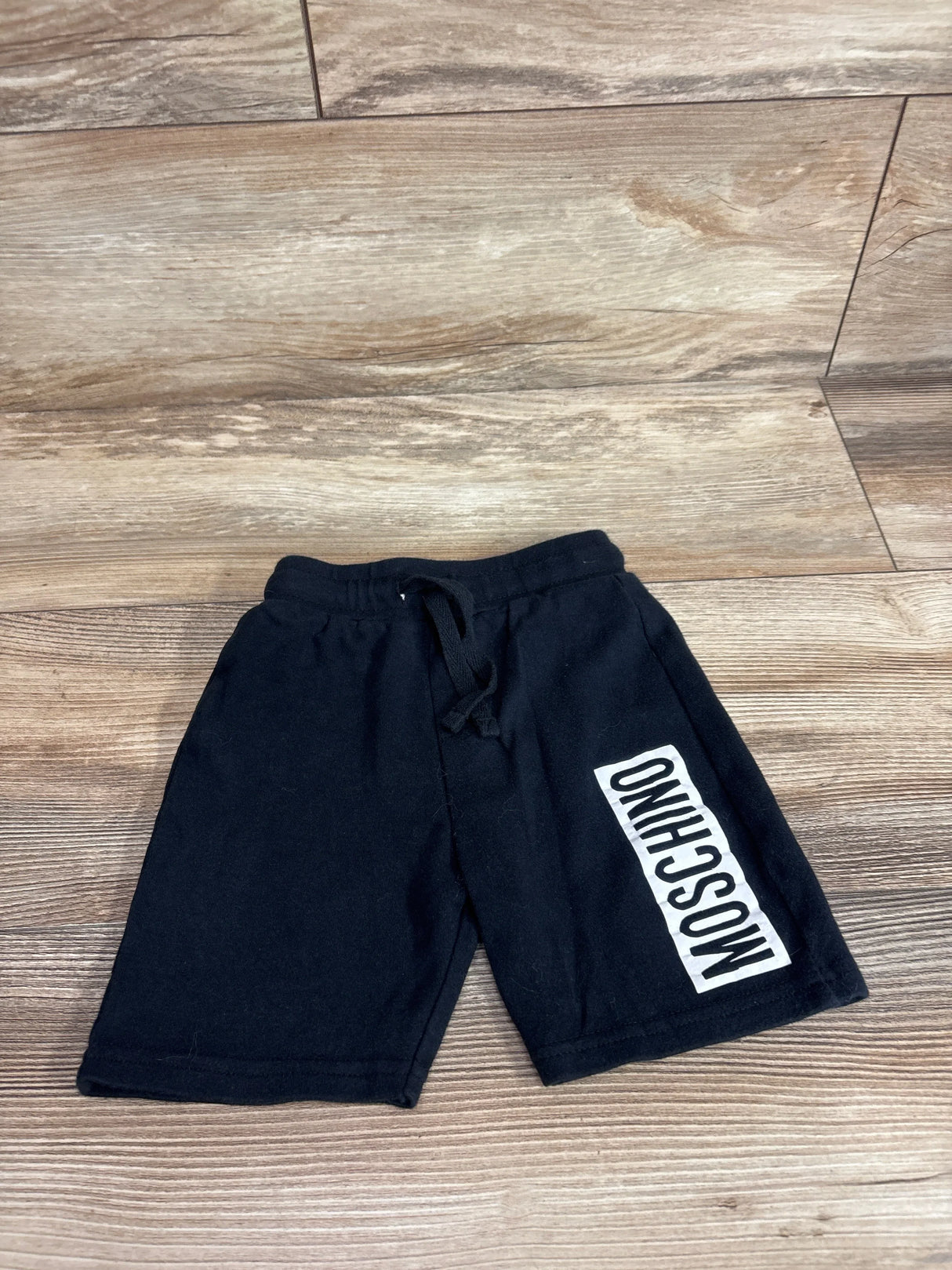 Flat lay of black Moschino baby shorts (12 months) showing vertical white MOSCHINO logo on left leg on wood floor background
