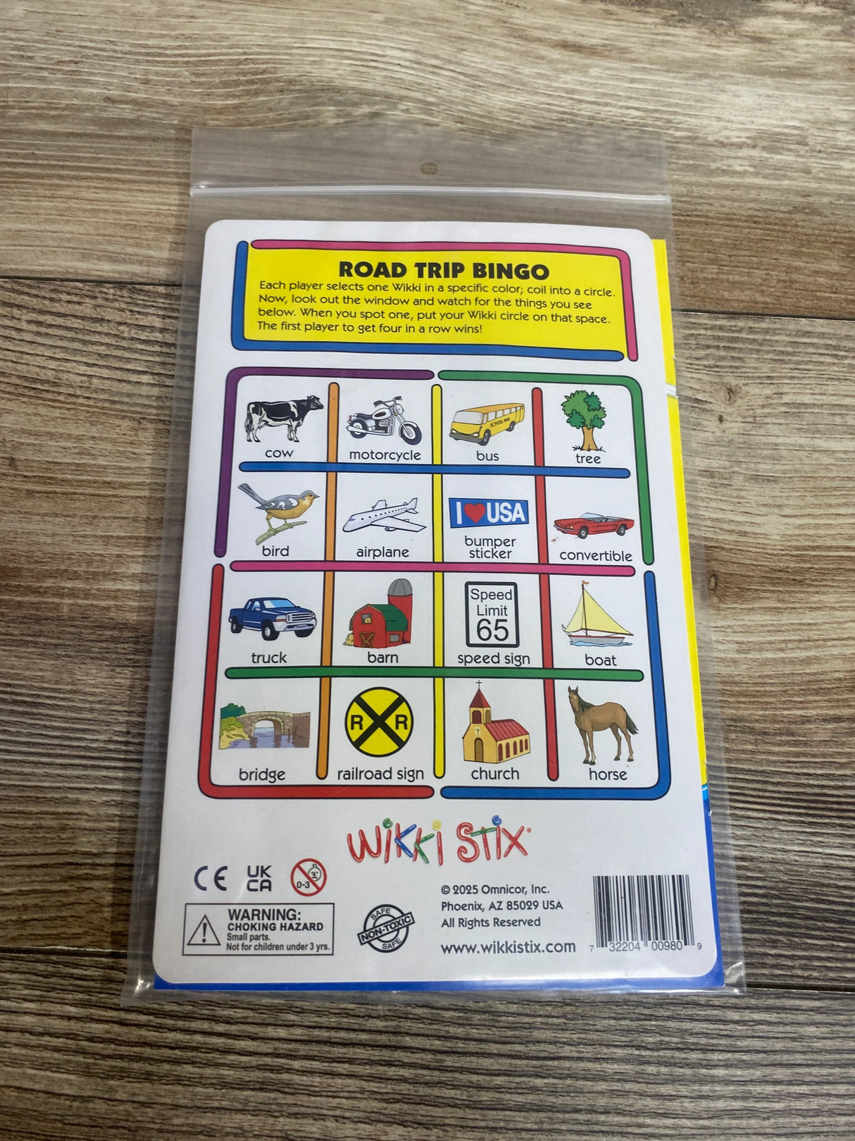 Wikki Stix Road Trip Bingo card with illustrations of cow, bus, motorcycle, airplane, and more on wooden surface