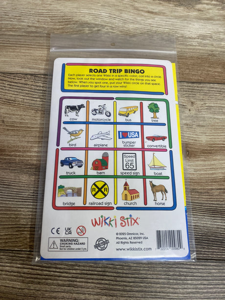 Wikki Stix Road Trip Bingo card with illustrations of cow, bus, motorcycle, airplane, and more on wooden surface