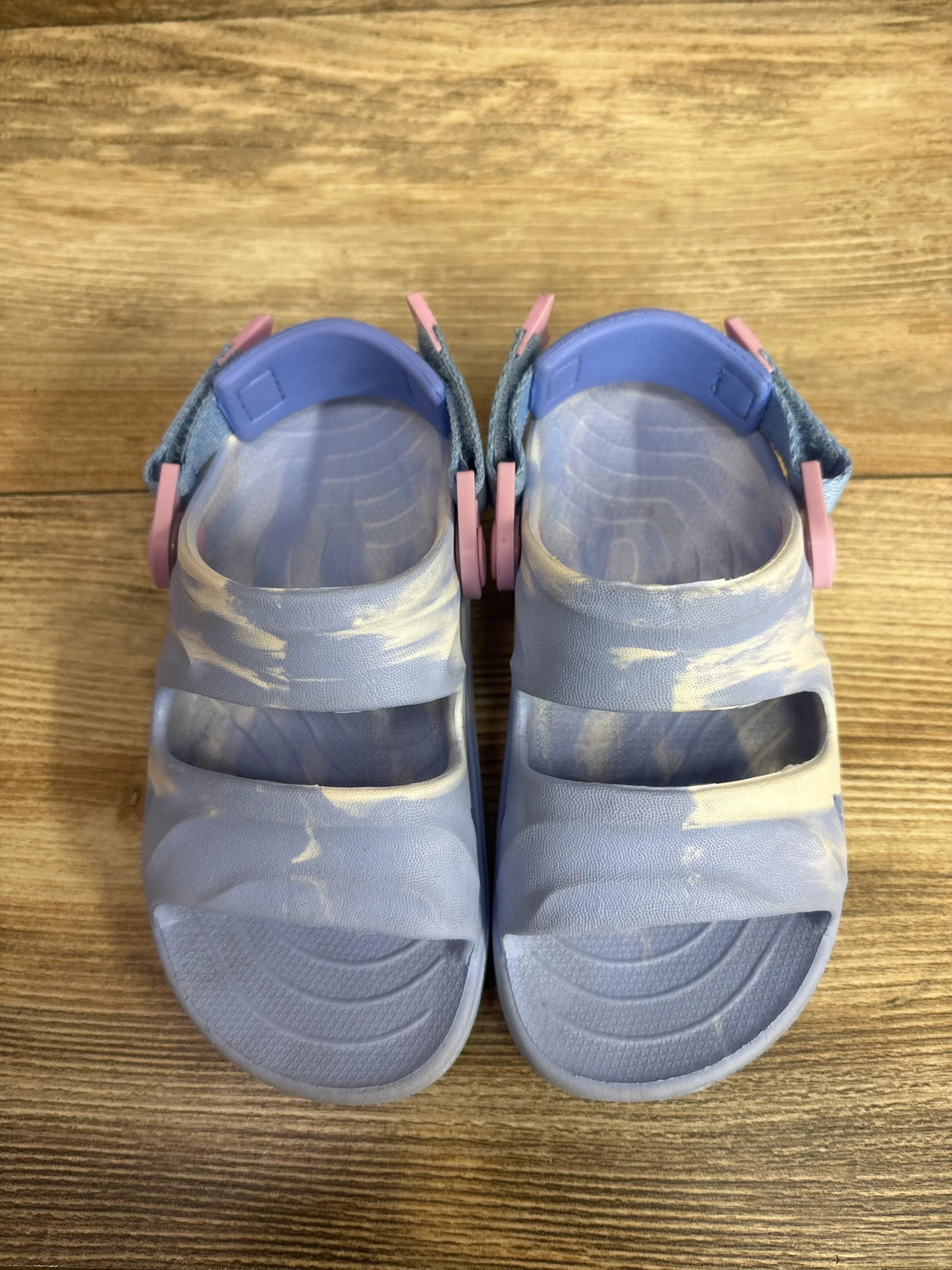 Overhead view of light blue Rugged Shark toddler sandals with marbled pattern, two straps and textured footbeds on wood