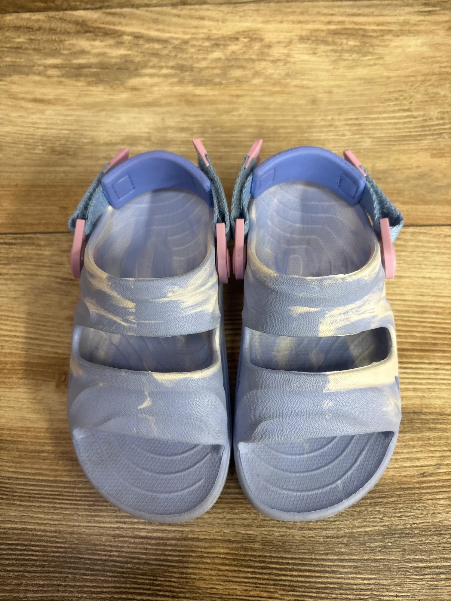 Overhead view of light blue Rugged Shark toddler sandals with marbled pattern, two straps and textured footbeds on wood