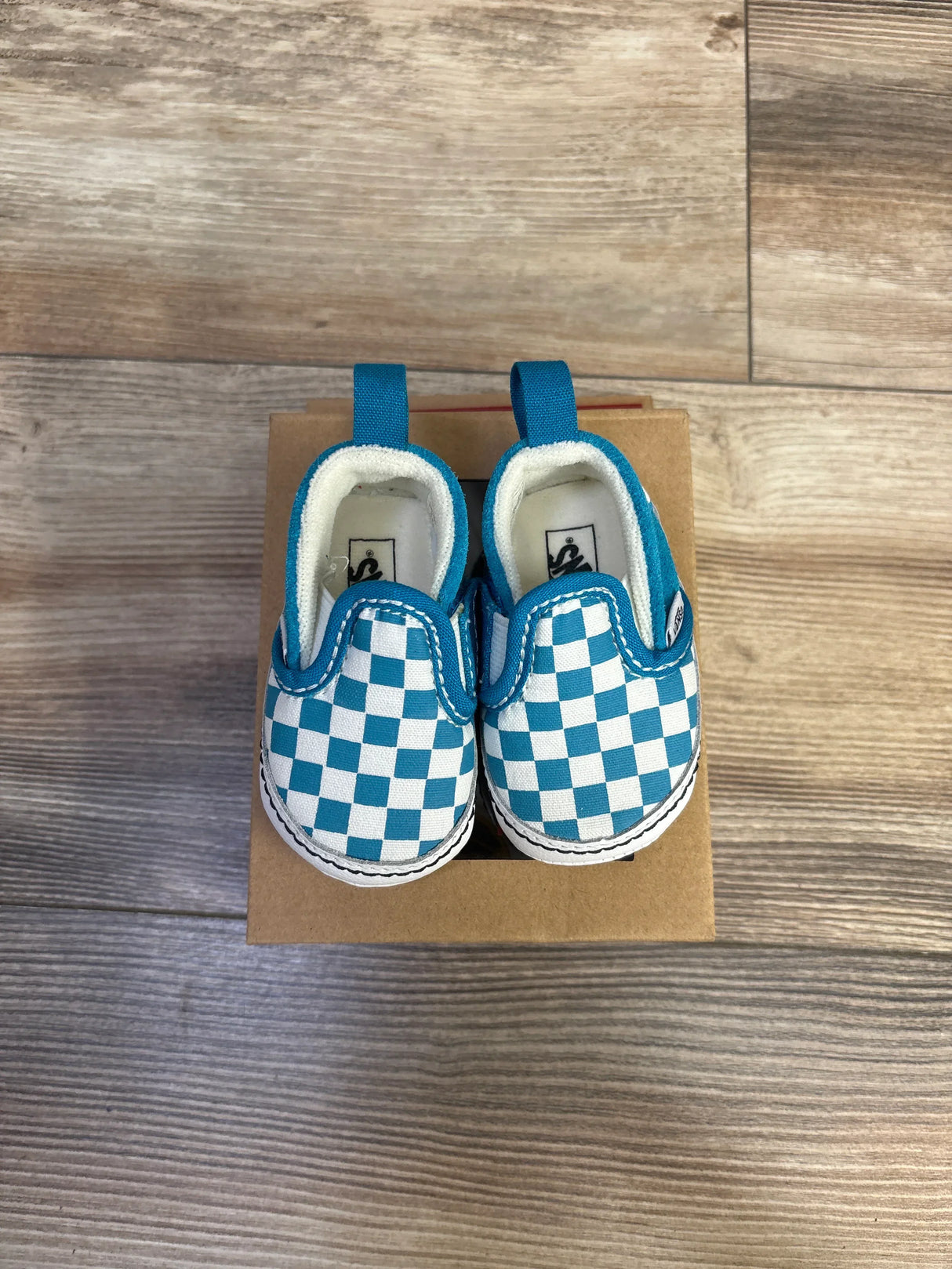Top-down view of Vans baby slip-on crib shoes showing blue-and-white checkered canvas and pull tabs on a small brown box on wood floor