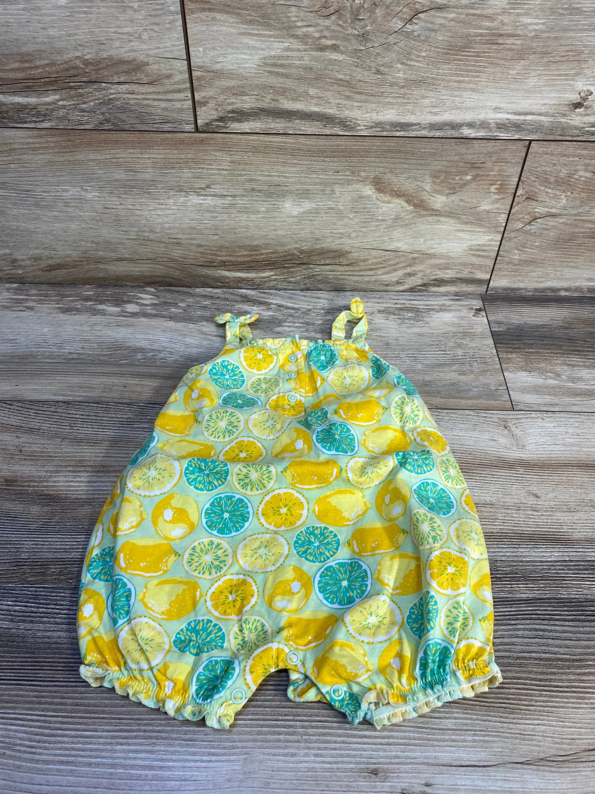 Top-down view of Henley lemon tank romper showcasing lemon print, knotted shoulder straps and snap crotch on wood floor