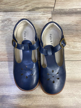 Top-down view of navy T-strap Mary Jane toddler flats showing flower-shaped toe cutouts and brass buckles on wood floor