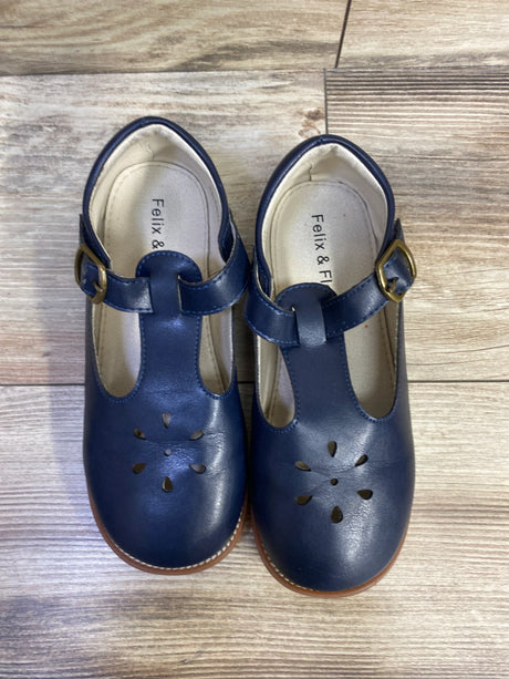 Top-down view of navy T-strap Mary Jane toddler flats showing flower-shaped toe cutouts and brass buckles on wood floor