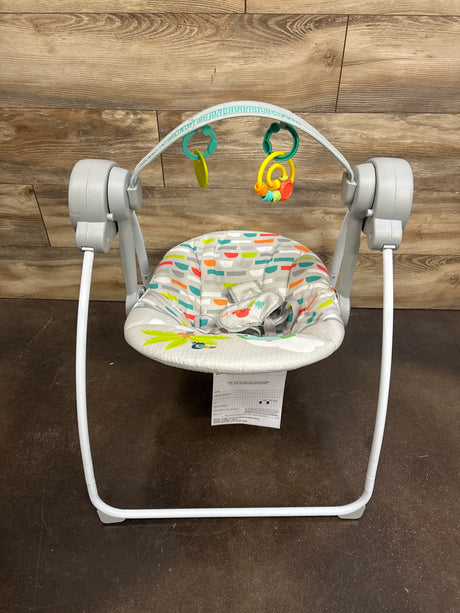 Front of Bright Starts portable compact baby swing with patterned seat, toy bar with two toys, harness, gray frame, wood wall