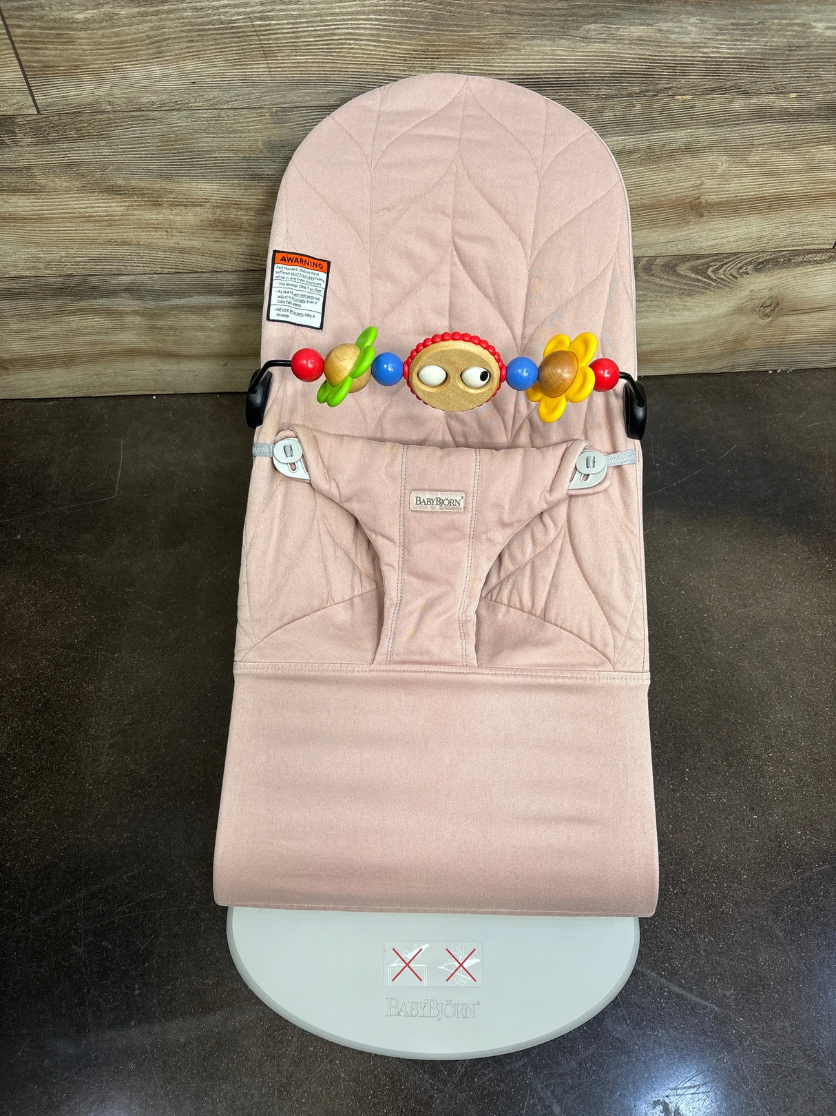 Overhead view of Baby Bjorn bouncer in dusty pink quilted petal fabric with colorful toy bar and front harness on wood floor