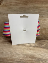 Back view of large striped hair bow showing horizontal rainbow stripes and metal clip on a white display card against wood background