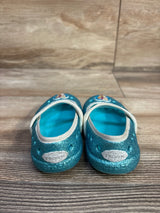 Front view of Crocs Frozen Mary Jane flats, size 5C, glittery teal blue showing white elastic strap and Frozen logo on toe with wood plank background