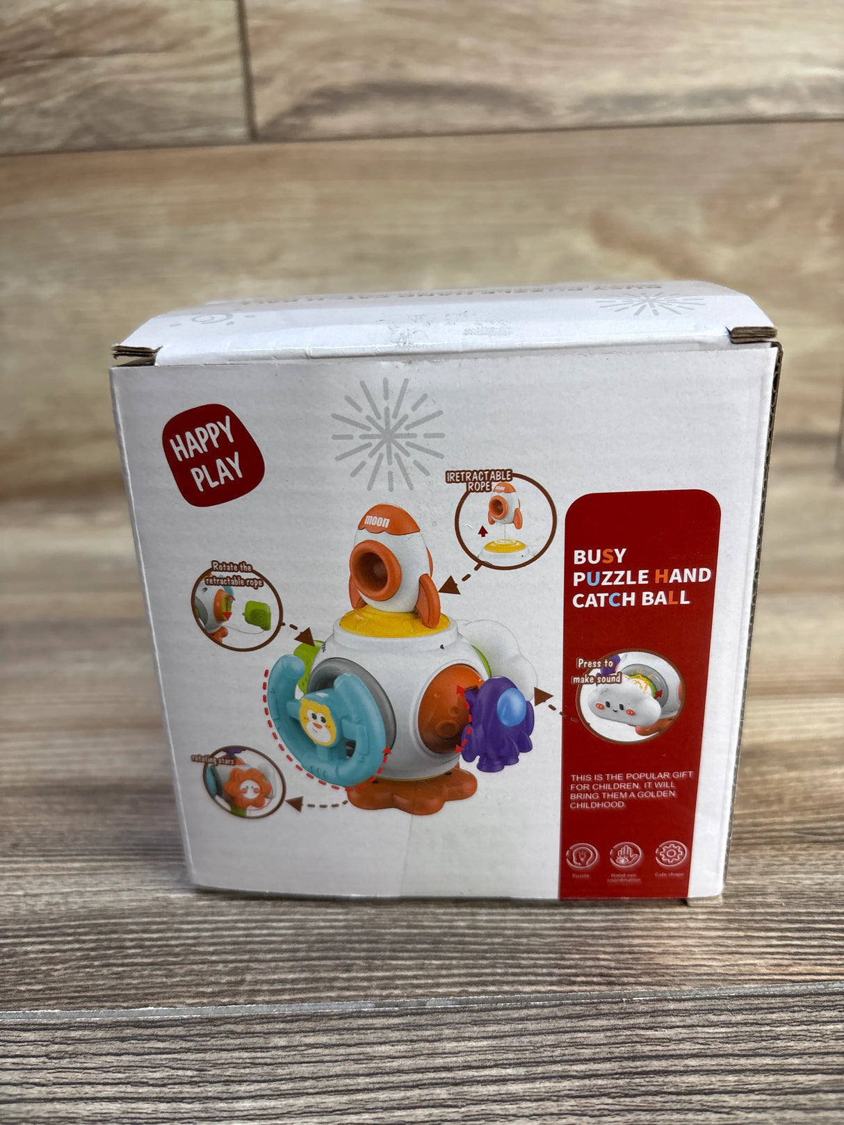 Front view of Happy Play Puzzle Busy Ball box with rotating puzzle panels and retractable rope on wooden floor