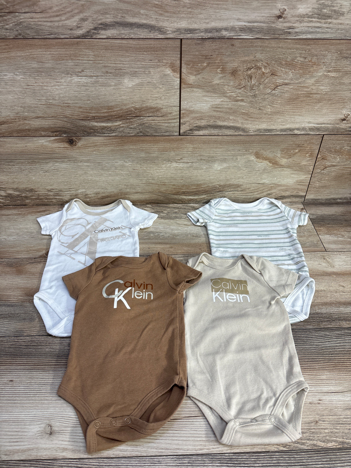 Four Calvin Klein branded baby bodysuits in white, brown, beige, and striped laid on wood floor