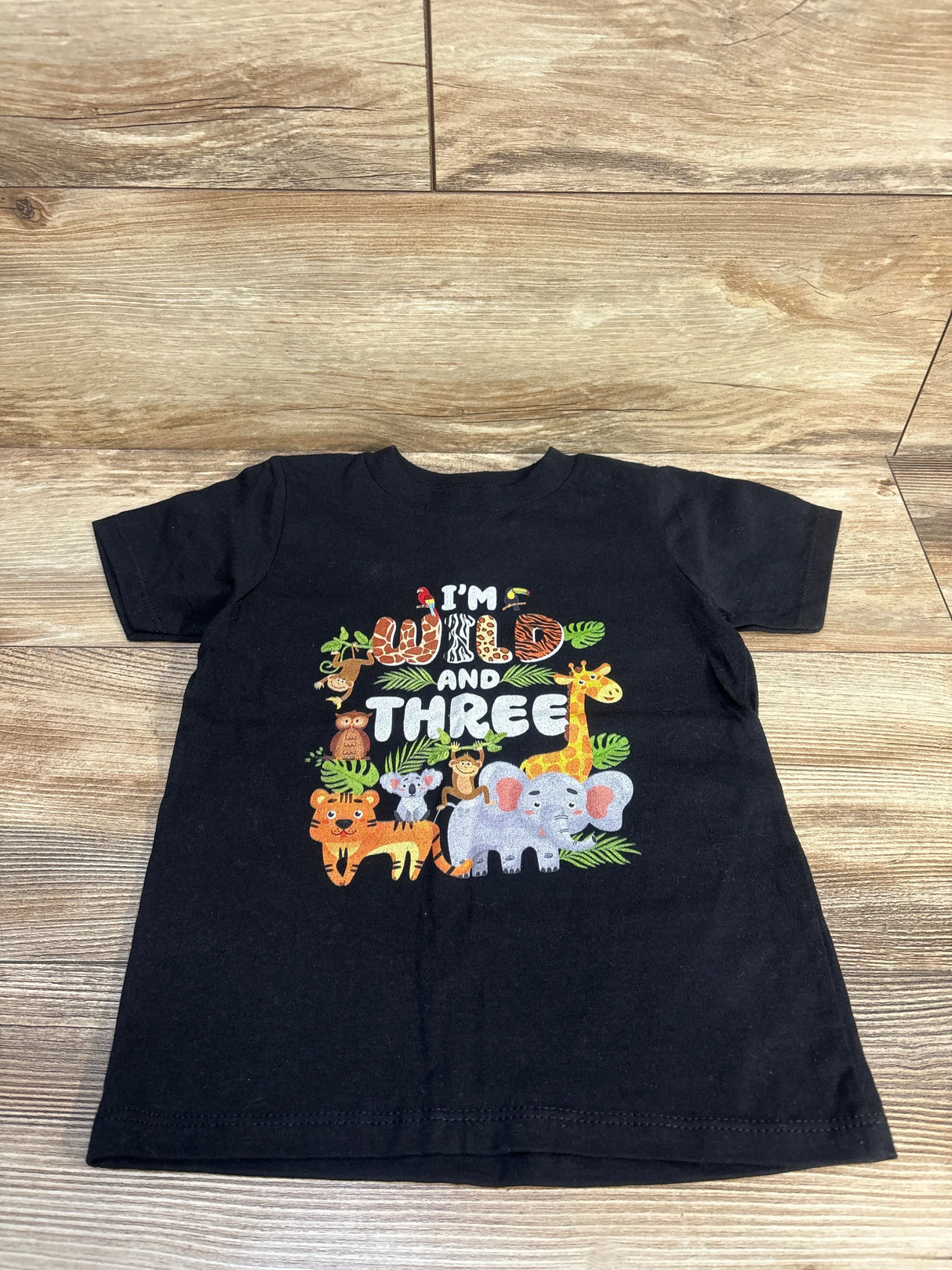 Front view of black Gildan toddler 4T t-shirt showcasing I'm Wild and Three jungle animal print on wood floor background
