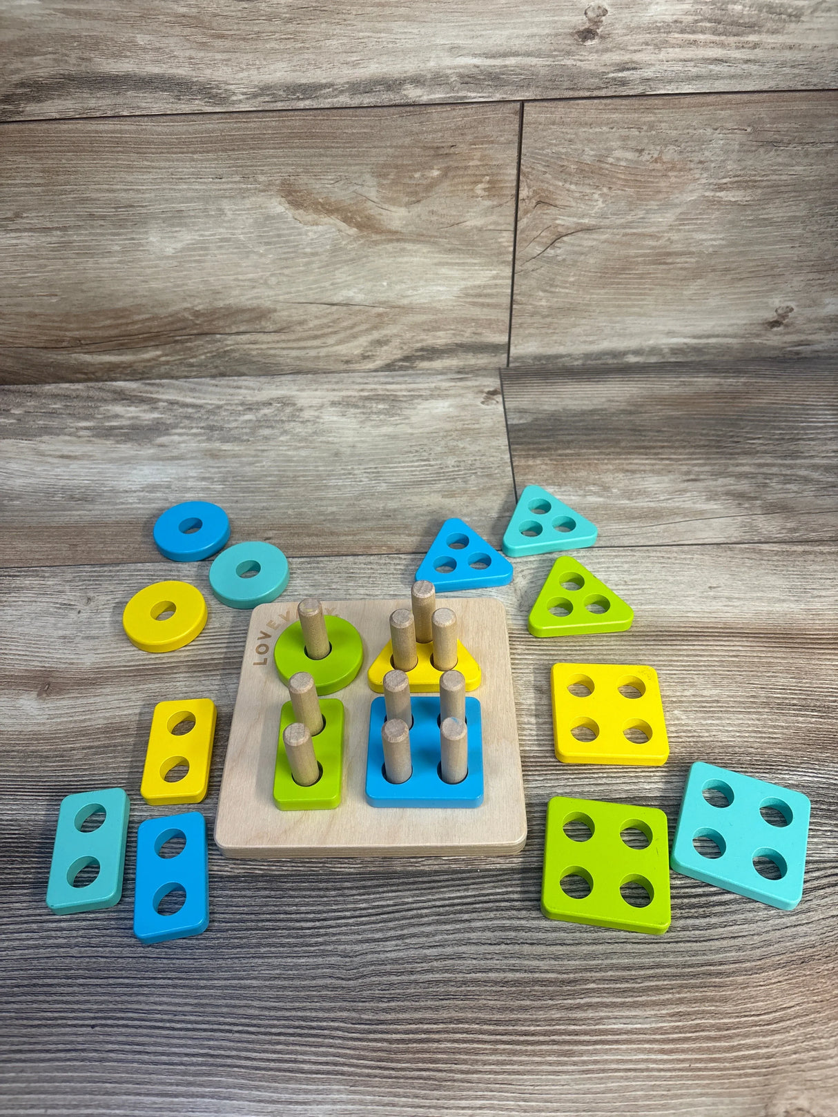 Top view of Lovevery Sort & Stack Peg Puzzle with wooden peg board and colorful geometric sorting pieces on a wood background