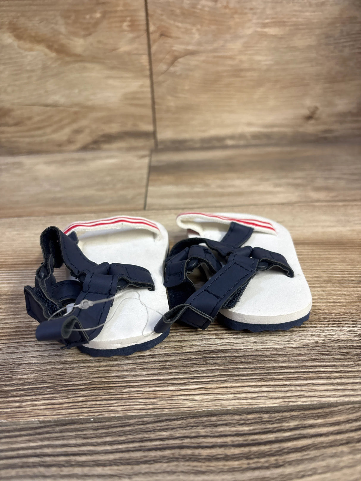 Front view of Child Of Mine baby sandals showing white footbed, navy velcro straps, red-striped heel on wood background