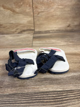 Front view of Child Of Mine baby sandals showing white footbed, navy velcro straps, red-striped heel on wood background