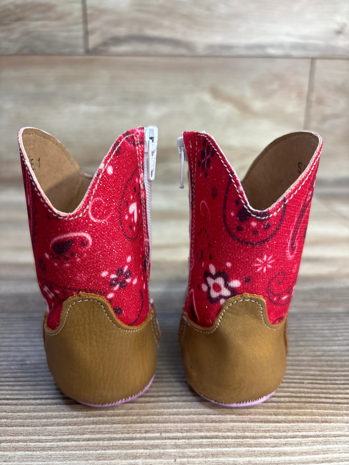 Rear view of Tin Haul Mini square-toe boots showing red paisley glitter shaft, tan soft sole heel and side zippers on wood floor