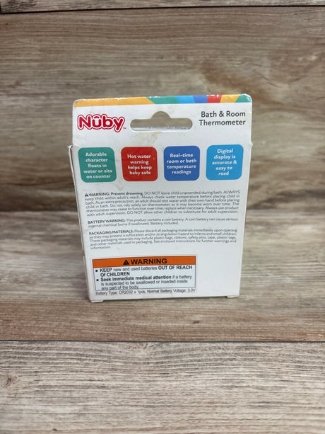 Back view of Nuby Bath & Room Thermometer packaging showing digital temperature display and hot-water warning on wood