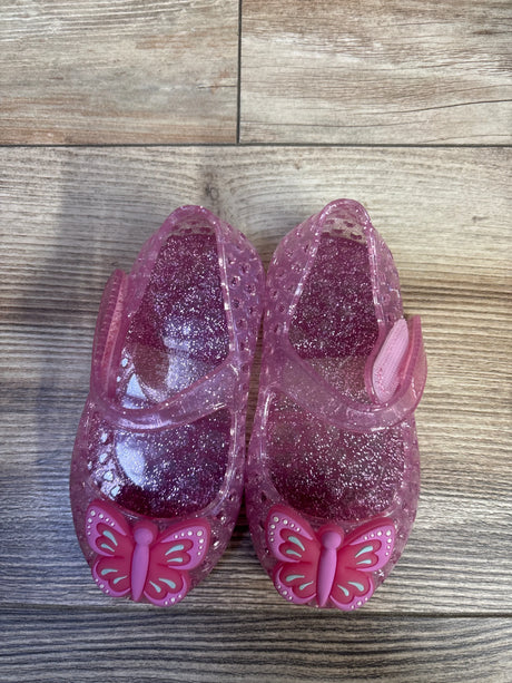 Top-down view of toddler pink glitter jelly Mary Jane flats showcasing butterfly toe charms and Velcro strap on wood floor